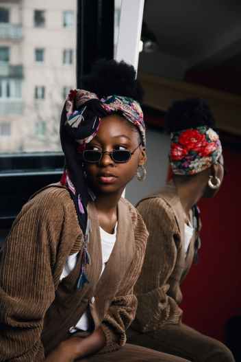 woman wearing white shirt and brown coat with sunglasses and floral turban taking selfie with mirror beside her